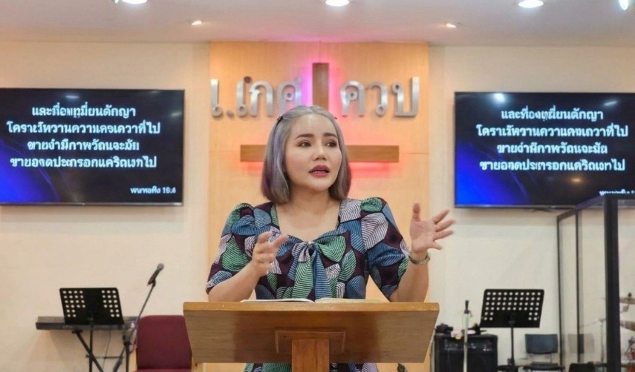 เรณู คารูโซ กำลังพูดบนเวที ถือพระคัมภีร์ที่เปิดอยู่ข้างกางเขนที่ส่องสว่าง